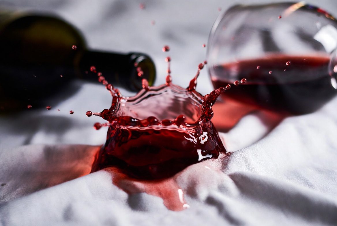SOS Mancha de Vinho: Vinho tinto atingindo uma camisa branca, demonstrando a urgência da limpeza.