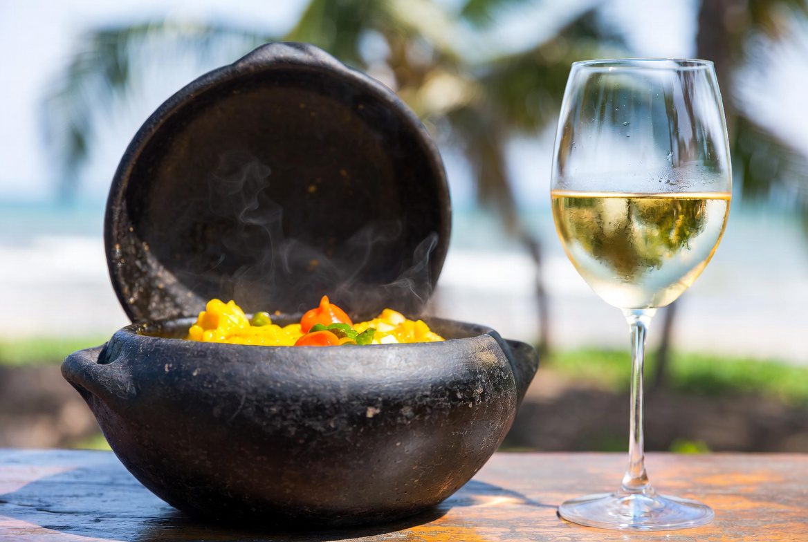 Vinho para Moqueca: Uma panela de barro com moqueca baiana ao lado de uma taça de vinho branco gelado.