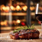 Um copo de vinho tinto ao lado de um prato de carne grelhada com alecrim em uma mesa de madeira rústica, ilustrando a harmonização perfeita.